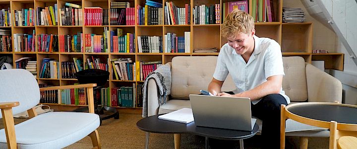 Glad videregåendeelev sitter på biblioteket med sin laptop og får eksamenshjelp online