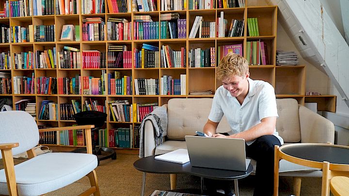 Glad videregåendeelev sitter på biblioteket med sin laptop og får eksamenshjelp online