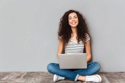 Smilende kvinne sitter med laptop-en på fanget på gulvet